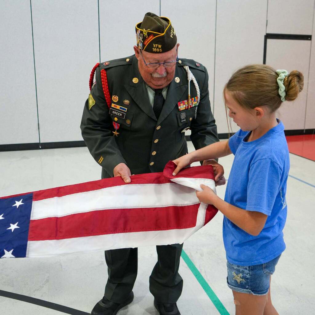 Image 7: A veteran in uniform carefully demonstrating the precise triangular fold technique to a young girl.