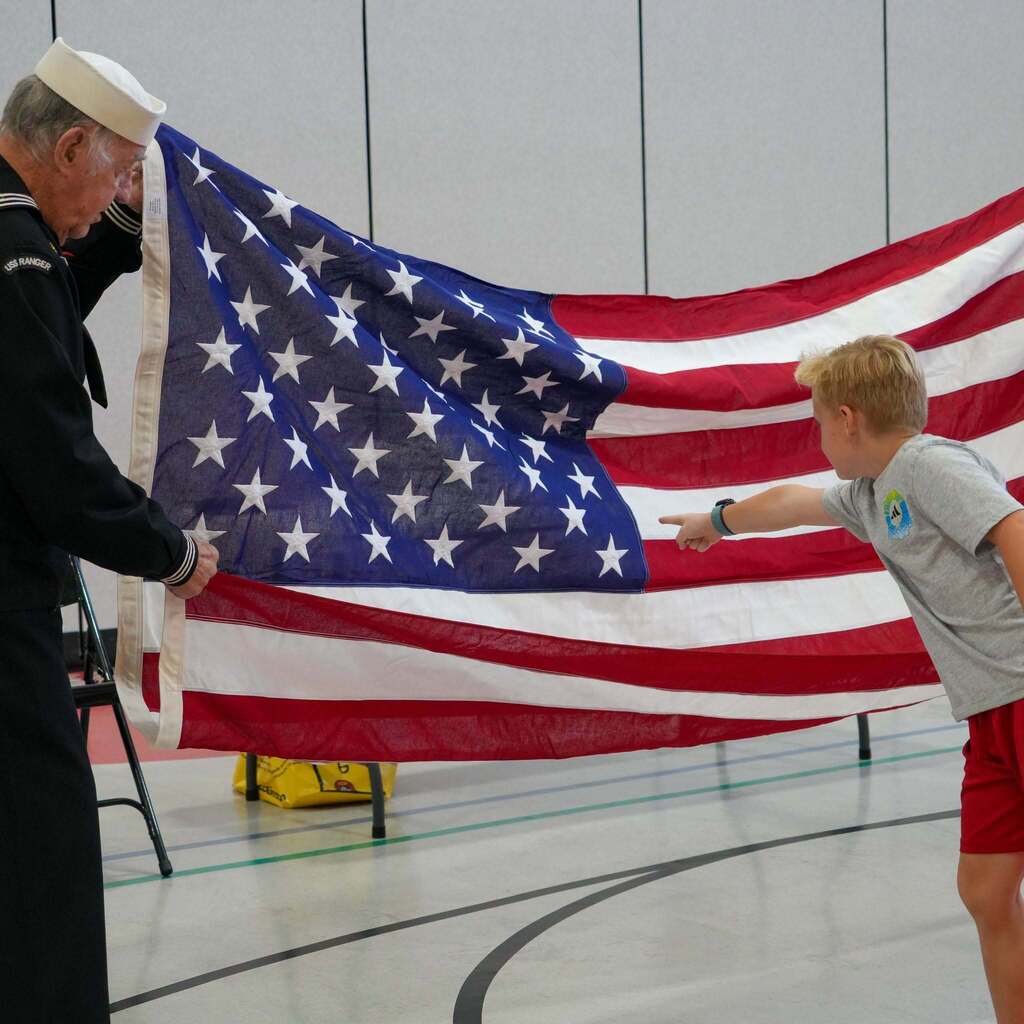 Image 4: A young student pointing toward the blue star field of an American flag held open by a Navy veteran in uniform.