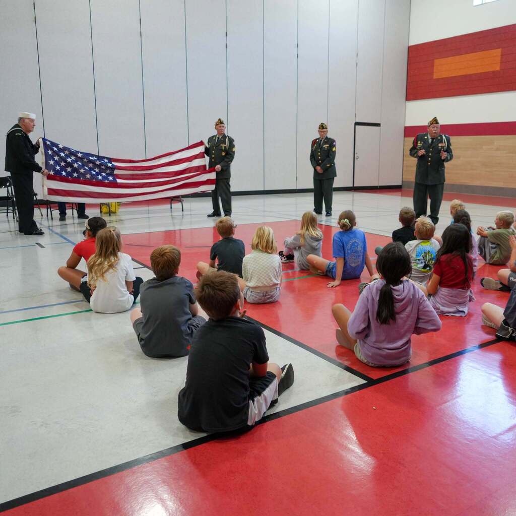 Image 3: A group of elementary students seated on the floor watching veterans hold up a fully unfurled American flag during a flag etiquette presentation.