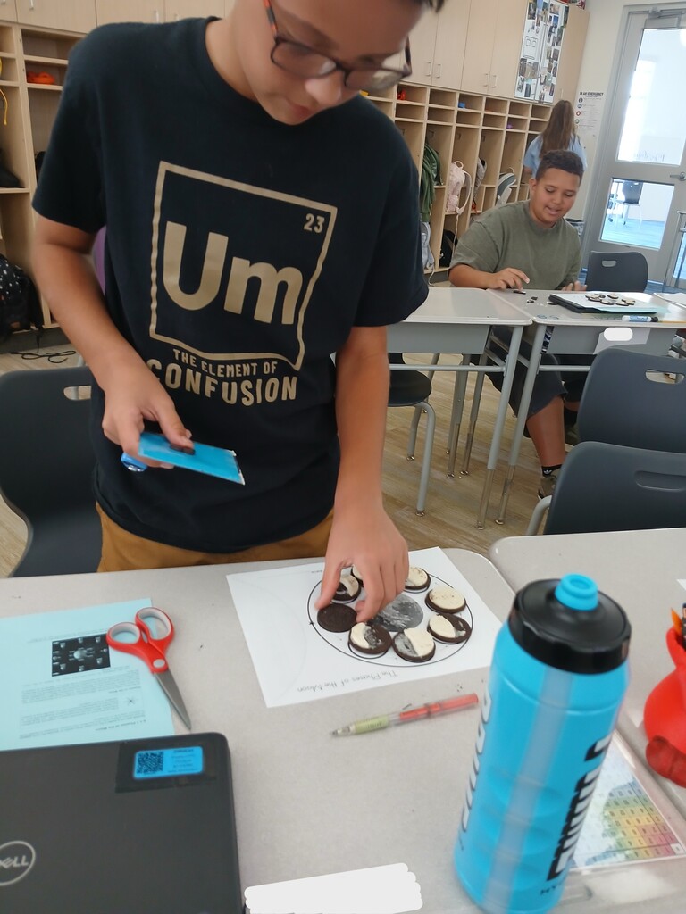 A student wearing a shirt reading "Um: The Element of Confusion" placing sandwich cookies onto a "The Phases of the Moon" diagram at a classroom desk.