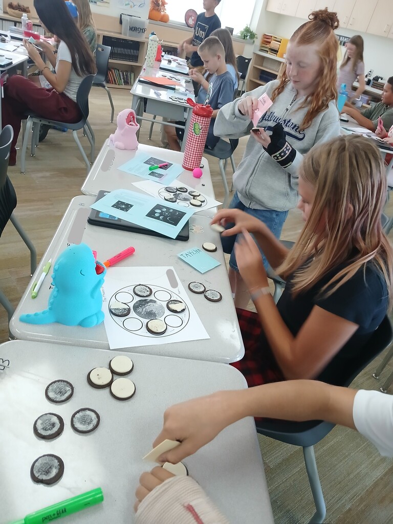 Students working at classroom desks on a moon phases activity using cookies, with one student wearing a "Yellowstone" sweatshirt.