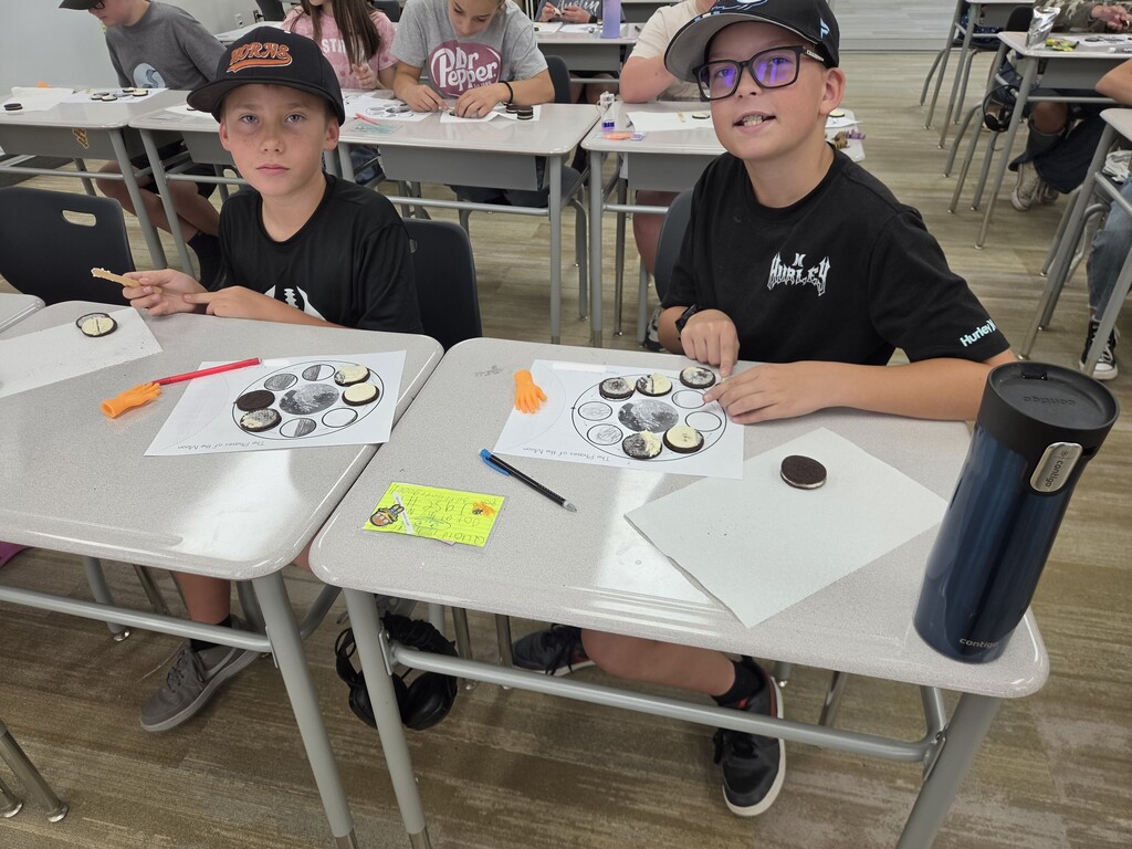 Two boys seated at classroom desks working on a "The Phases of the Moon" cookie activity, with one wearing a hat reading "HORNS" and a shirt featuring "Hurley".