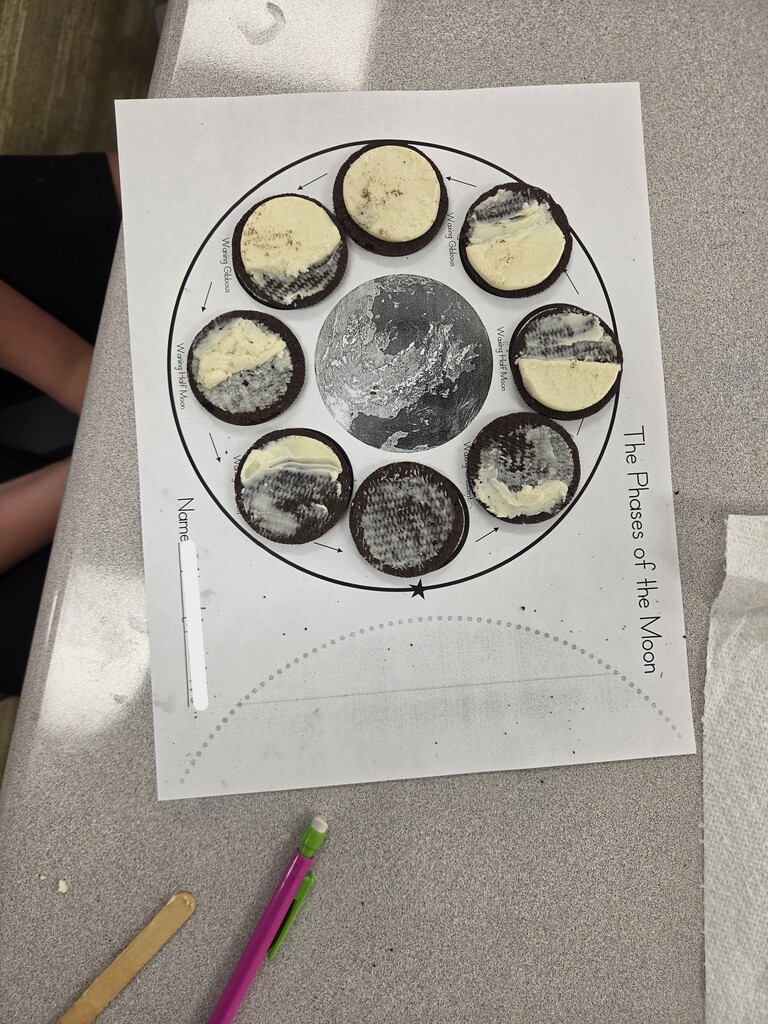 Top-down view of a student paper titled "The Phases of the Moon" with scraped cream sandwich cookies representing moon phases around Earth, labeled with terms like "Waxing Gibbous", "Waxing Half Moon", and "Waning Half Moon".