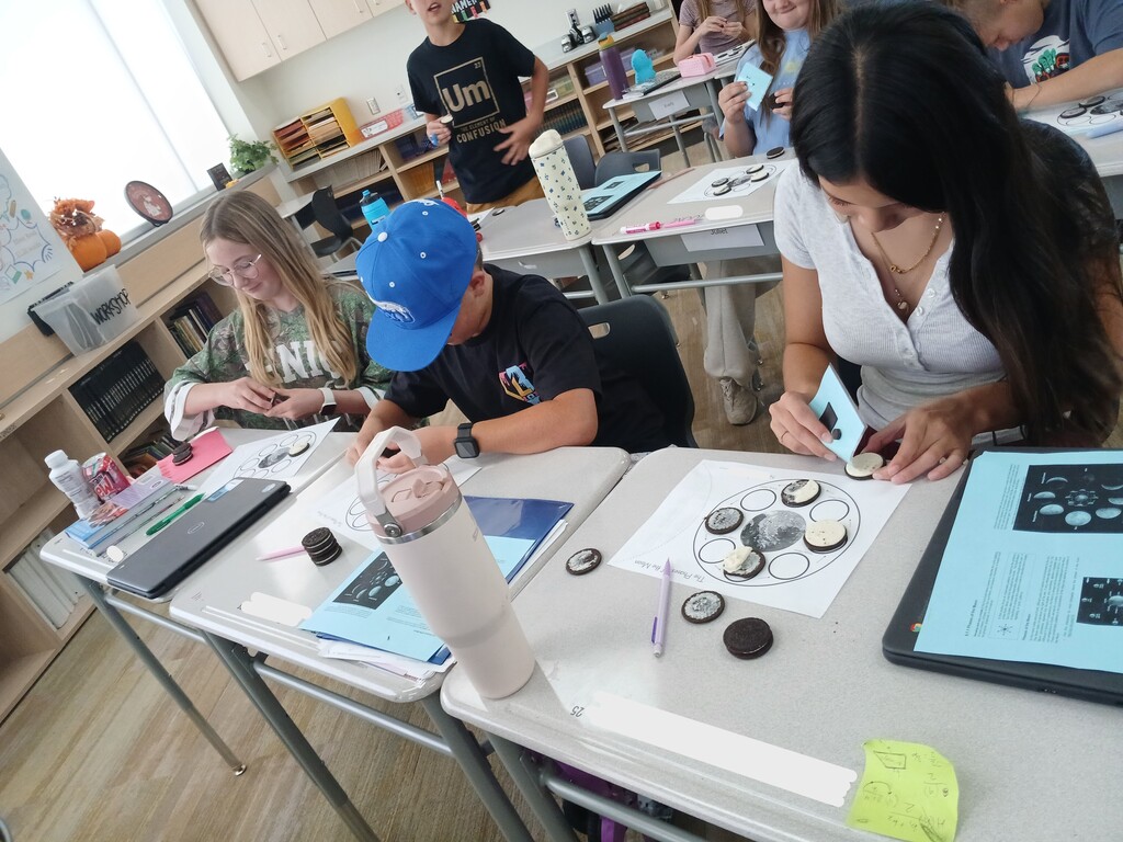 Students seated at classroom desks working on a "The Phases of the Moon" cookie diagram, with a student in the background wearing an "Um: The Element of Confusion" t-shirt.