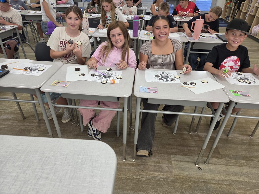Four students smiling at classroom desks while arranging sandwich cookies on a science worksheet titled "The Phases of the Moon"