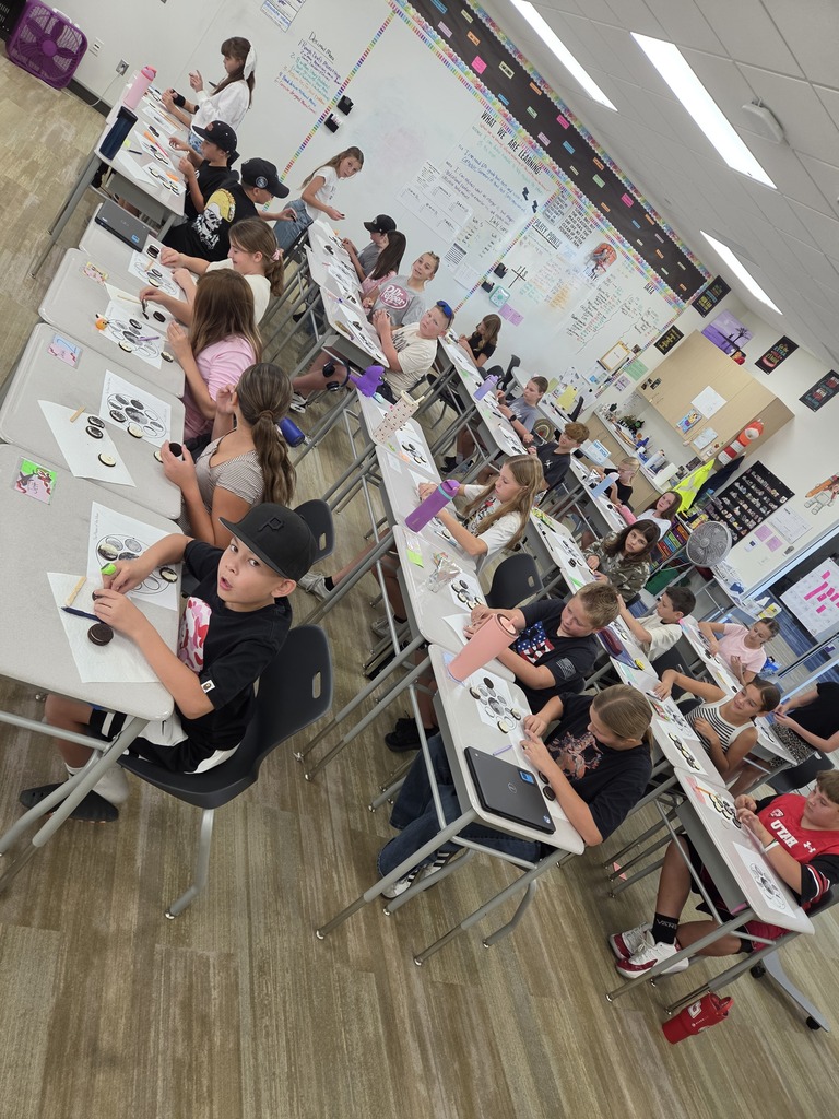 High-angle view of a classroom full of students seated at rows of desks creating moon phase diagrams using cookies, with a whiteboard in the background titled "WHAT WE ARE LEARNING"