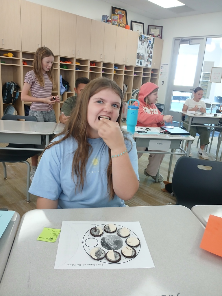 A girl eating a cookie at her classroom desk sitting behind a completed "The Phases of the Moon" worksheet.