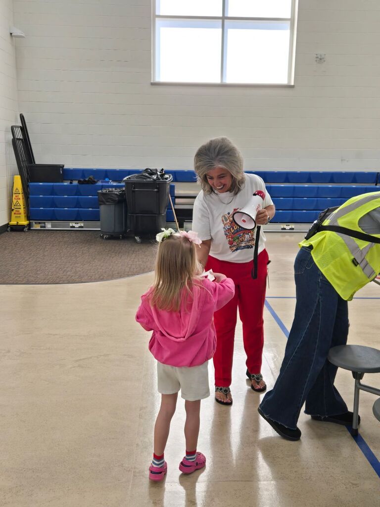 A woman holding a Pyle megaphone speaks with a young girl in a pink jacket near a yellow caution cone labeled "CAUTION WET FLOOR" and "CUIDADO PISO MOJADO".