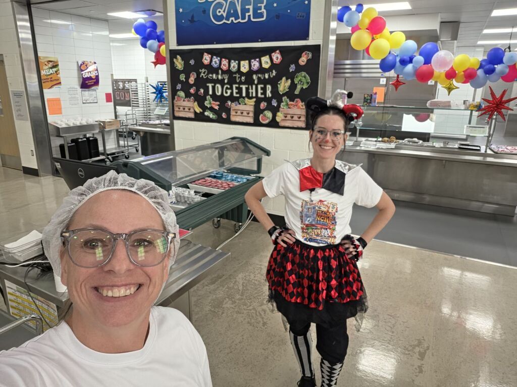 A cafeteria staff member in a hairnet and a colleague in a jester costume stand in front of a banner reading "Ready to grow TOGETHER" under a sign reading "CAFÉ" with posters reading "MEAT," "GRAINS," and "NATIONAL SCHOOL LUNCH WEEK" visible nearby.
