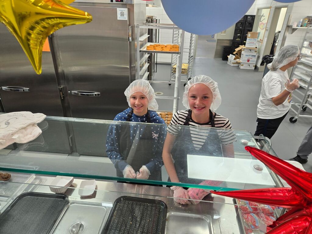 Two young girls wearing hairnets and aprons smile behind a glass cafeteria serving counter decorated with star balloons, with boxes labeled "FRUIT LAND APPLES" stacked in the background.