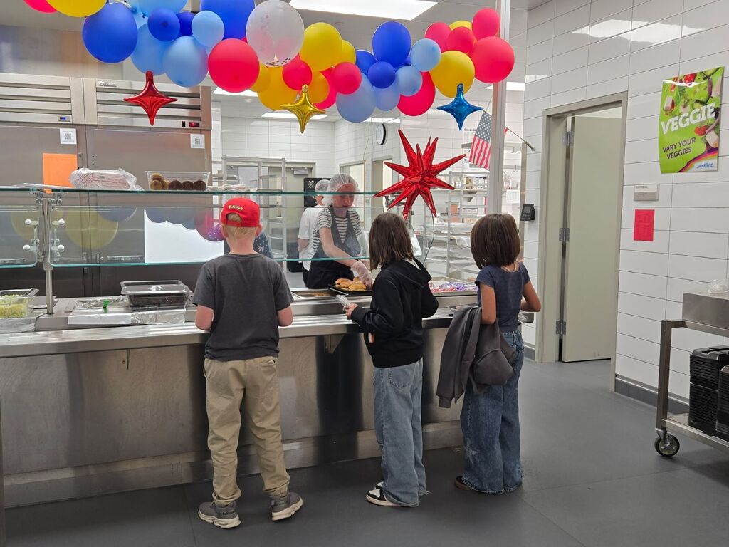 Three students stand in a cafeteria line decorated with colorful balloons and star decorations near a poster that reads "VEGGIE - VARY YOUR VEGGIES".