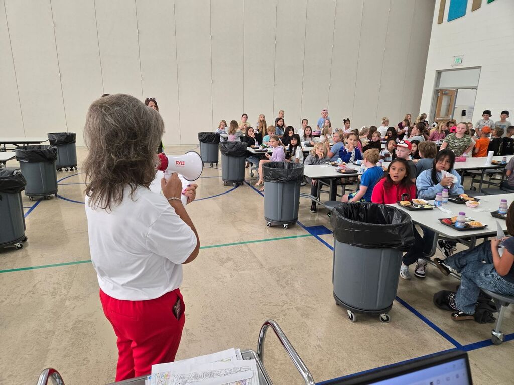 A woman with a red Pyle megaphone addresses a group of elementary school students sitting at cafeteria tables.