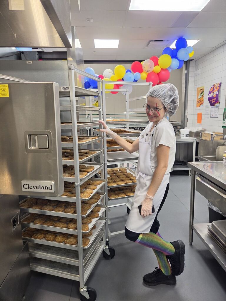 A cafeteria worker in a hairnet, apron, and clown face paint poses near a Cleveland-branded heating cabinet filled with food trays under colorful balloons and wall signs reading "MEAT" and "GRAINS".