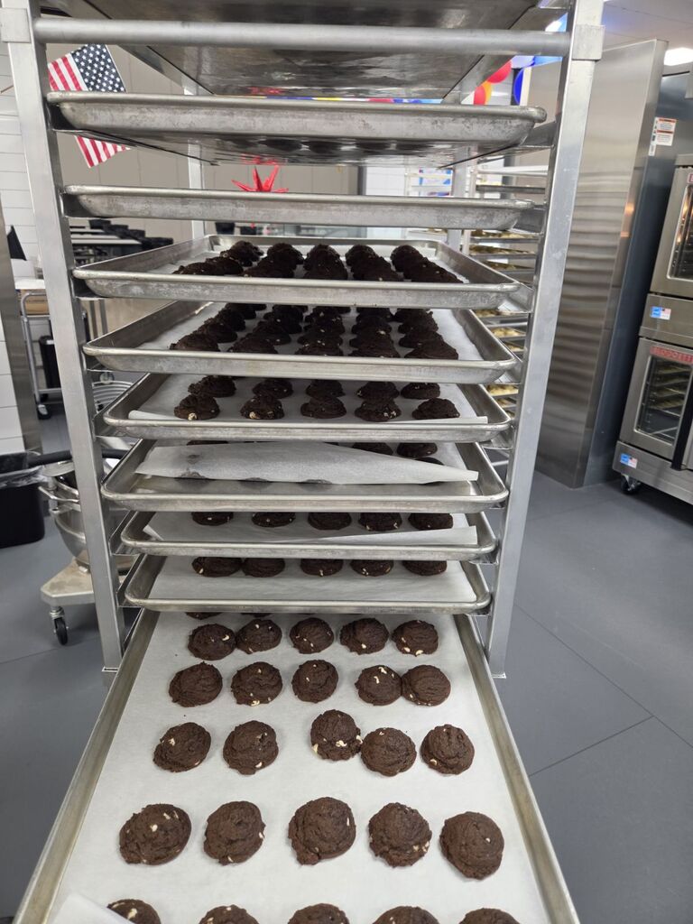 A tall metal bakery rack filled with sheet pans containing freshly baked chocolate cookies, positioned in front of an American flag.