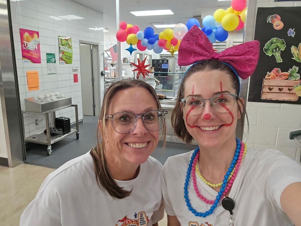 Two school cafeteria staff members wearing festive outfits and clown makeup stand together in front of wall signs reading "FRUIT" and "VEGGIE".