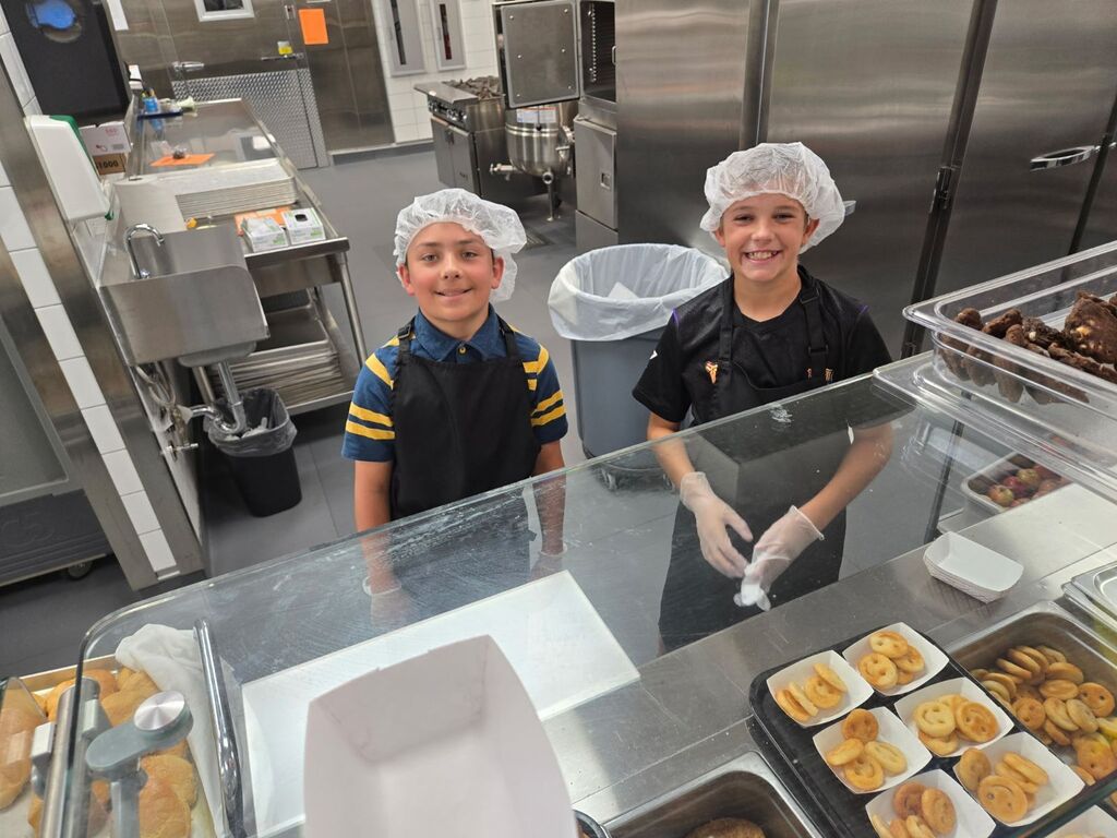 Two smiling boys wearing hairnets, aprons, and gloves stand behind a cafeteria serving counter filled with smiley-face potato fries and cookies.