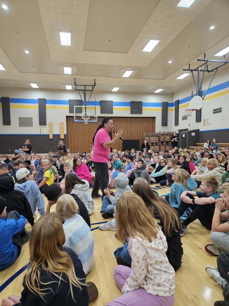 A wide view of the Lomond View gymnasium filled with elementary students seated on the floor listening intently to storyteller Nestor in a bright pink shirt. 