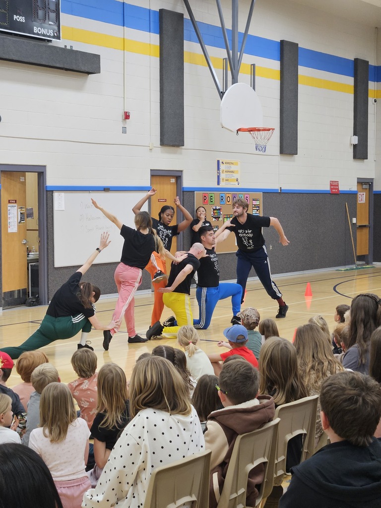 Dance troupe holding a synchronized pose in a line across the gym court floor in front of seated students.