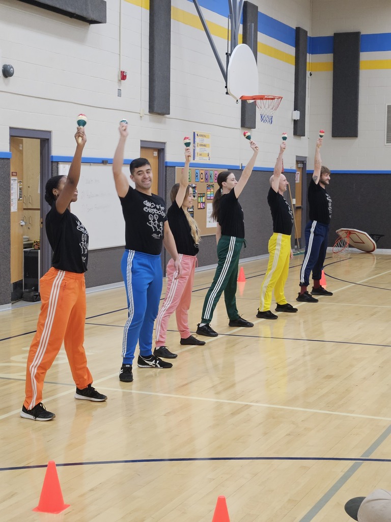 Six dancers in colorful track pants synchronized in a line holding maracas in the air.