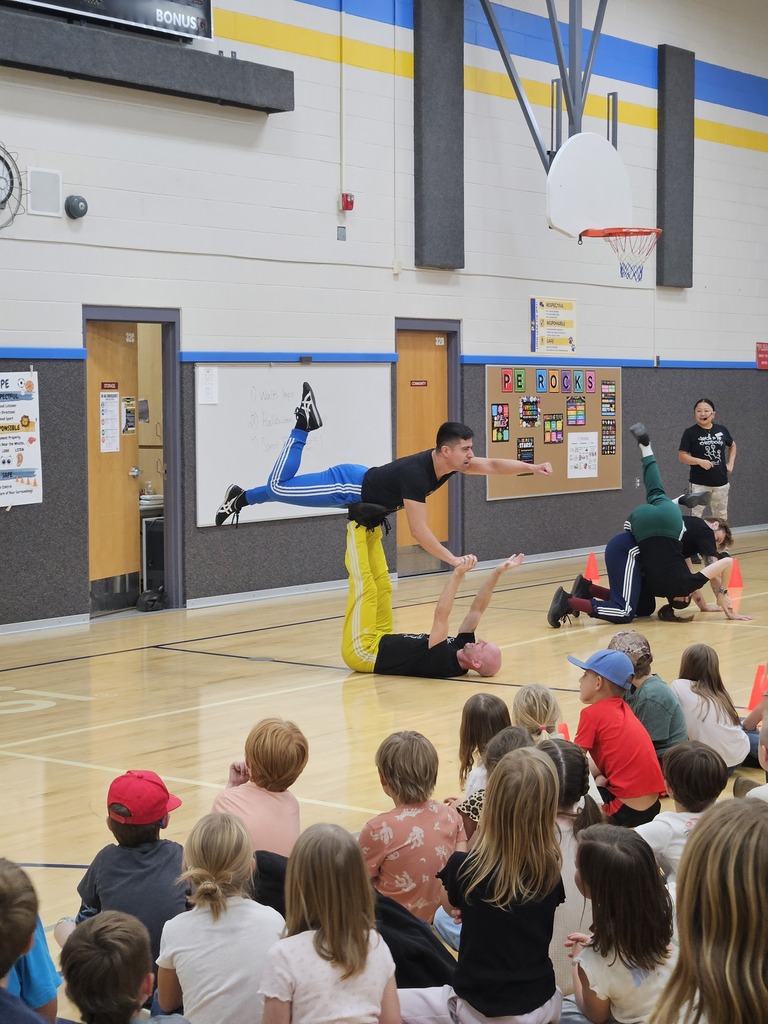 Dancers showcasing an impressive acrobatic balance move on the gym floor while elementary students watch from the audience.