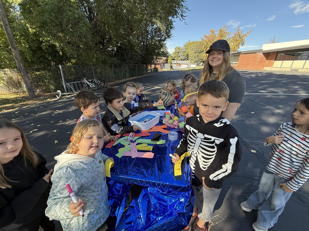 A group of kindergarten students and an adult volunteer standing around a blue outdoor table covered in colorful paper strips for Unity Day activities. 