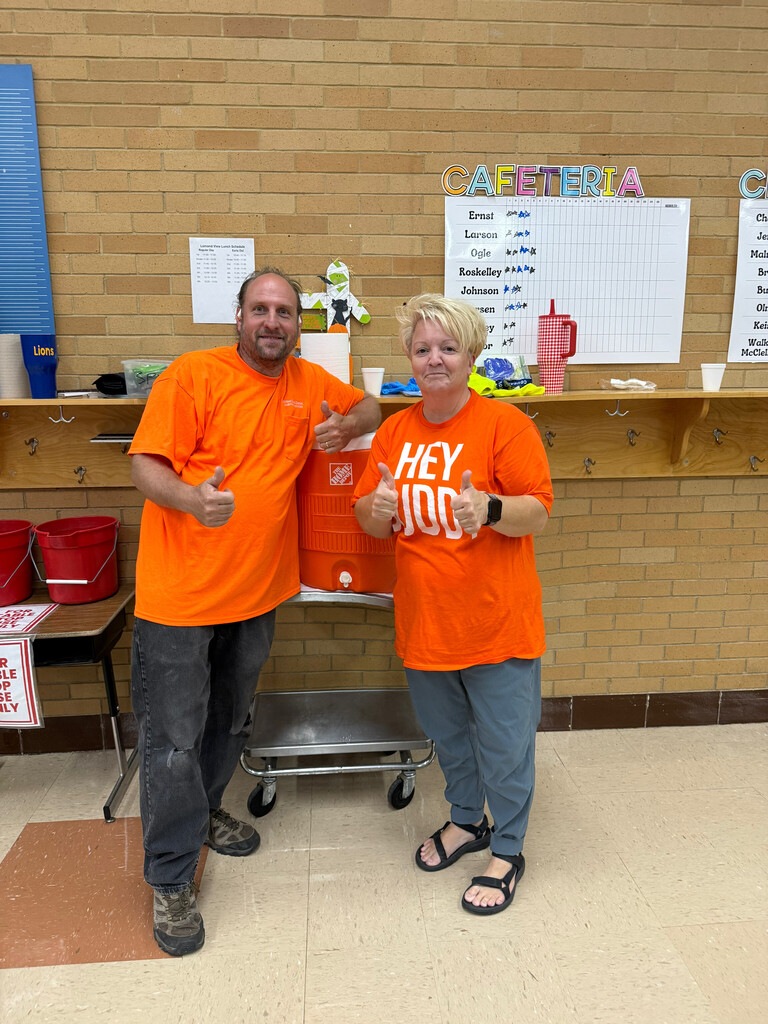 Miss Sara and Mr. Louis giving thumbs-up gestures while wearing bright orange shirts inside the school cafeteria.