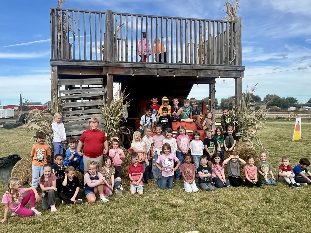A large group of first-grade students posing together in front of a rustic wooden play structure and vintage red truck outdoors.   