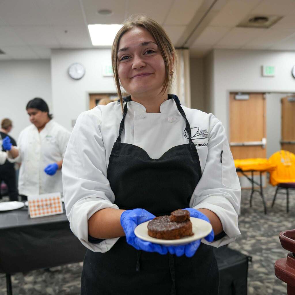 A smiling female culinary student wearing a white chef coat, black apron, and blue protective gloves, holding out a small plate with a freshly baked chocolate dessert