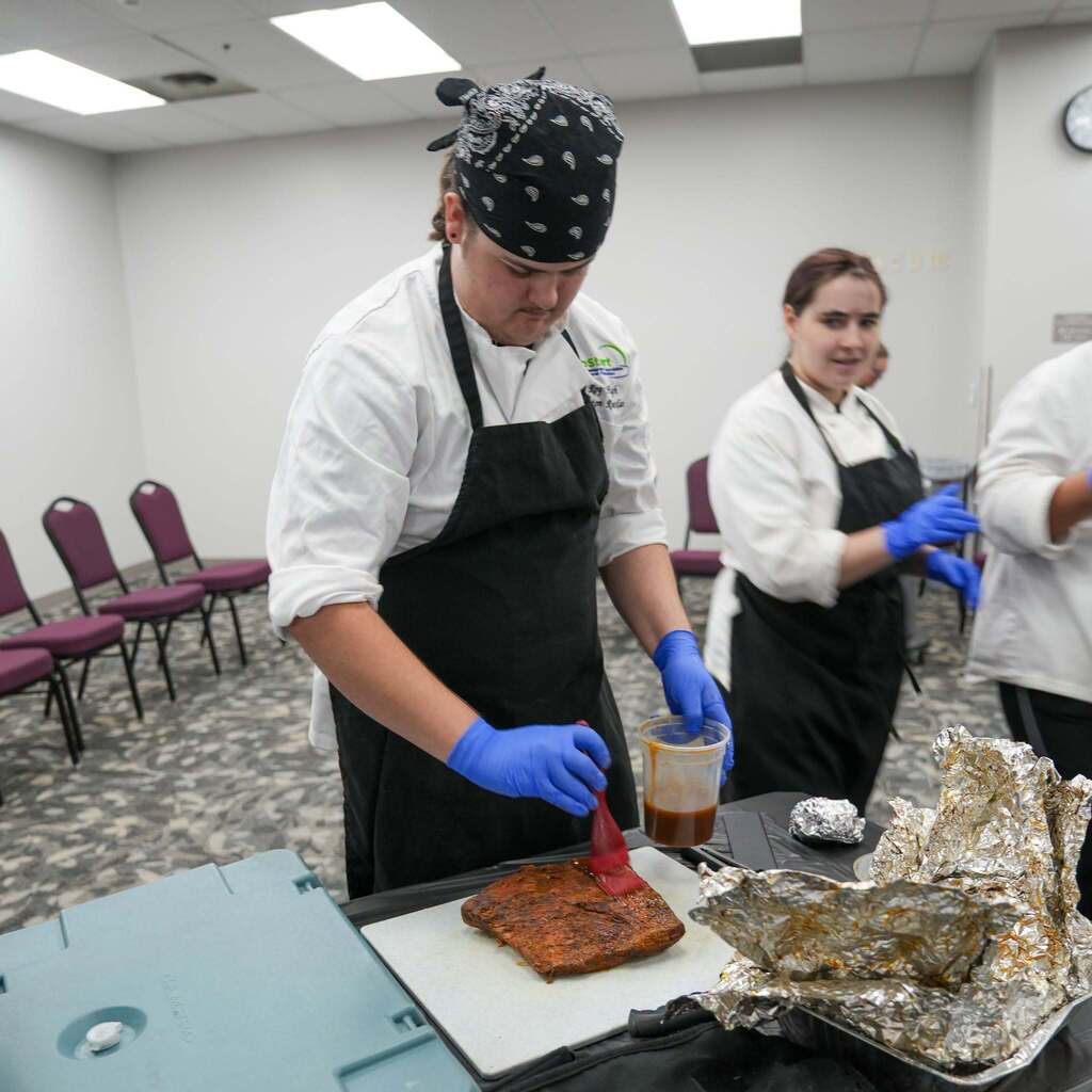 A high school culinary student in a black bandana, chef coat, and blue gloves using a pastry brush to apply sauce to a piece of meat on a cutting board. Another student in the background works nearby.