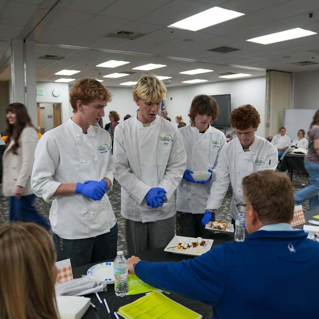 Four high school culinary students in white chef coats presenting plated dishes to judges seated across a table. The judges inspect the food entries and evaluation sheets.