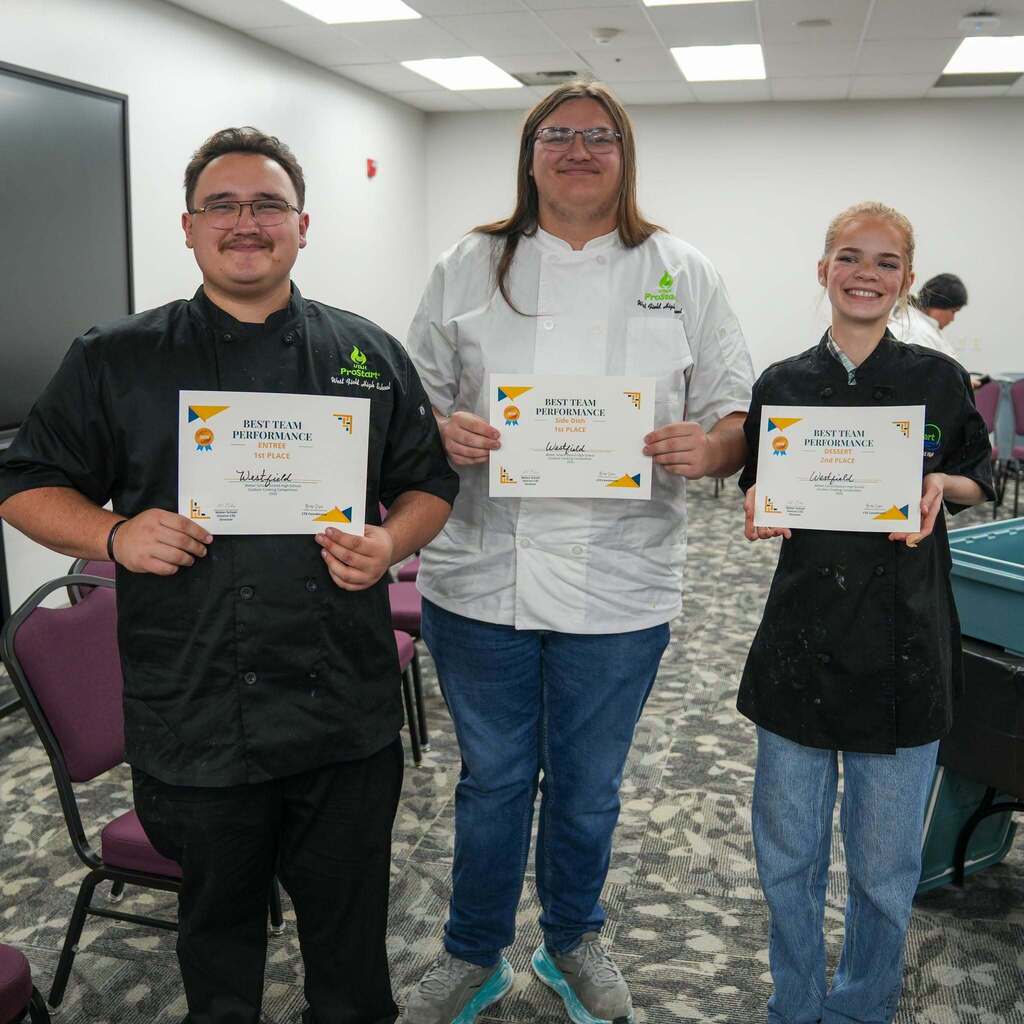 Three high school culinary students standing together in a room, smiling and holding certificates from a ProStart culinary competition. The students are wearing white chef coats or black aprons. The certificates read "BEST TEAM PERFORMANCE" for Entree 1st Place, Side Dish 1st Place, and Dessert 2nd Place.