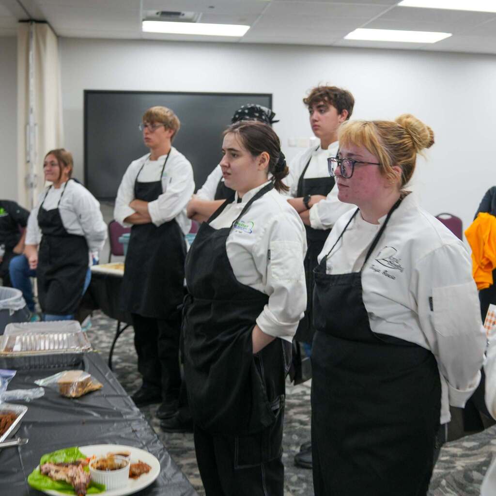 A lineup of culinary students wearing white chef coats and black aprons standing attentively in a row while waiting for their dishes to be evaluated at a competition table.