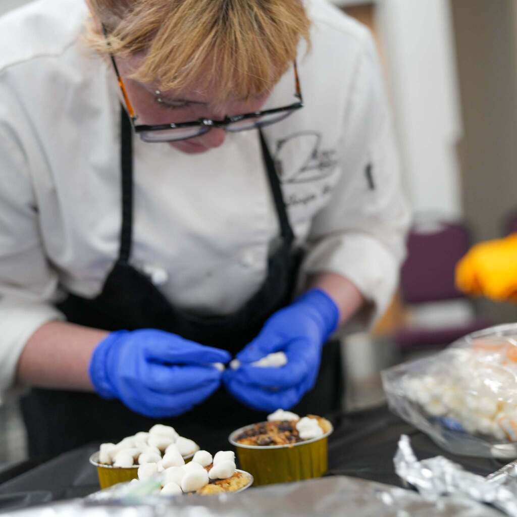 Close-up of a student chef in glasses and blue gloves meticulously placing mini marshmallows on top of individual baked desserts in gold tins.