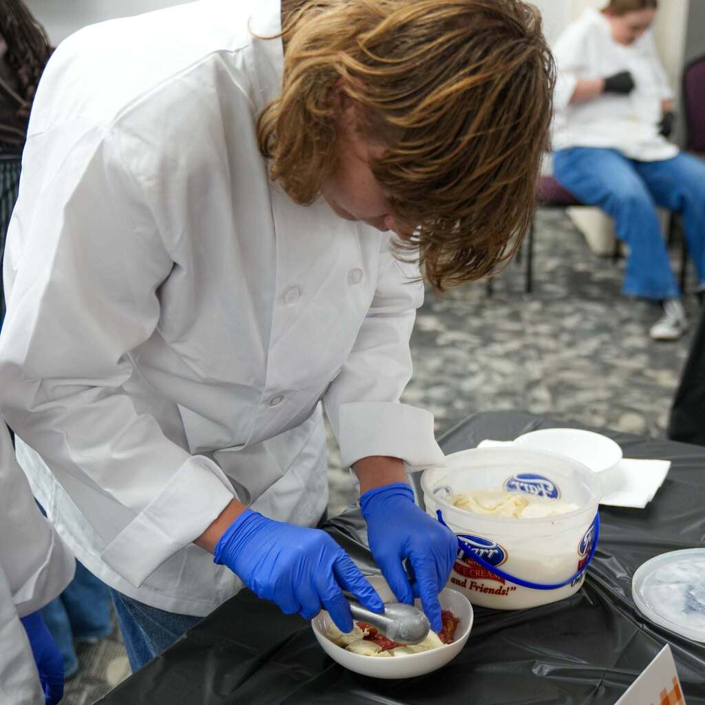Close-up of a student in a white chef coat and blue gloves carefully scooping vanilla ice cream from a bucket into a small serving dish over a dessert.