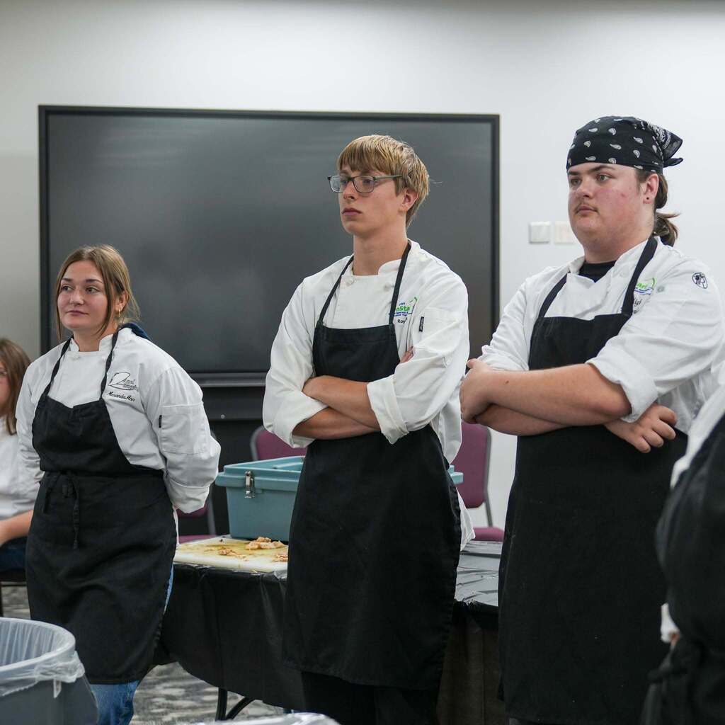 Three high school culinary students standing side-by-side with arms folded or at their sides, listening attentively during a competition evaluation. They are wearing white chef coats and black aprons.