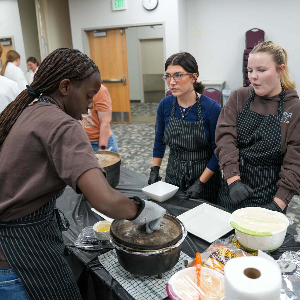 Three culinary students in striped aprons gathering around a table during a cooking competition. One student with braided hair reaches into a Dutch oven with a protective glove, while the other two look on attentively.