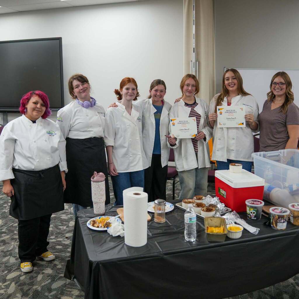 A group of six culinary students and an adult instructor standing behind a food display table holding certificates that read "BEST TEAM PERFORMANCE". The table is set up with paper towels, serving dishes, a red cooler, and food containers.