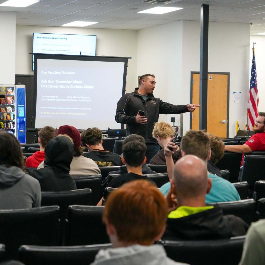 Image 7: A presenter pointing toward a projection screen displaying "One Next Step This Week: Ask Your Counselor About One Career You're Curious About" in front of a seated audience of students.