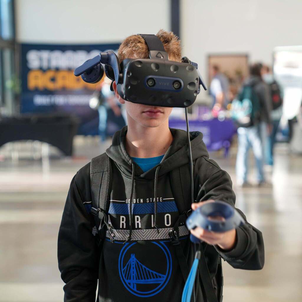 Image 1: A high school student wearing a Golden State Warriors hoodie and a Virtual Reality (VR) headset, holding a VR controller directly toward the camera during a hands-on CTE technology display.