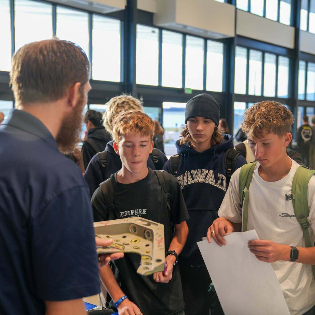 Image 3: A group of high school students listening intently as a CTE instructor shows them a custom-machined metal part during a career showcase event.