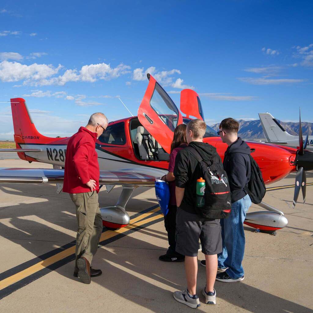 Image 10: A group of students gathered around a bright red and white single-engine airplane with gull-wing doors open on a sunny tarmac while an instructor explains features of the aircraft.