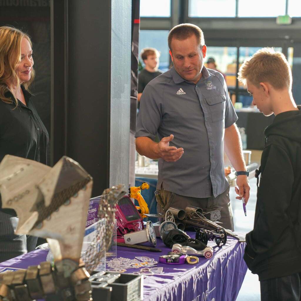 Image 2: An instructor in a grey shirt demonstrating precision manufacturing and engineering model cars and tools on a table covered with a purple cloth to an interested student.