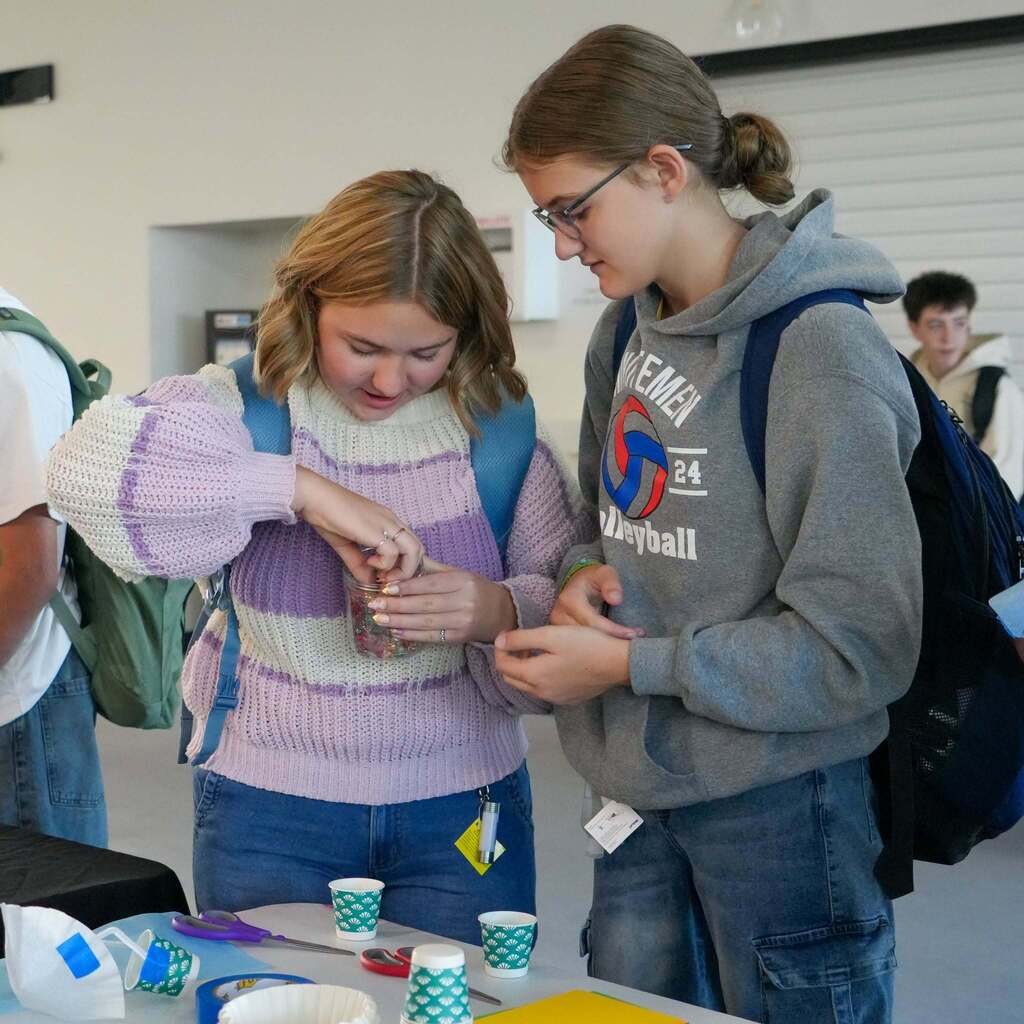 Image 5: Two female students collaborating on a hands-on activity with small craft cups, beads, and materials at a project booth.