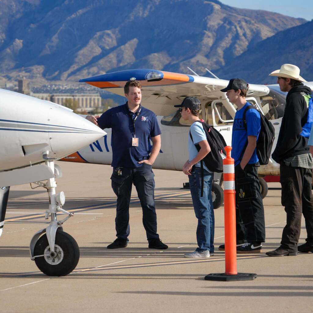 Image 8: An aviation instructor standing outdoors on the tarmac in front of a light aircraft, discussing flight components with three high school students.