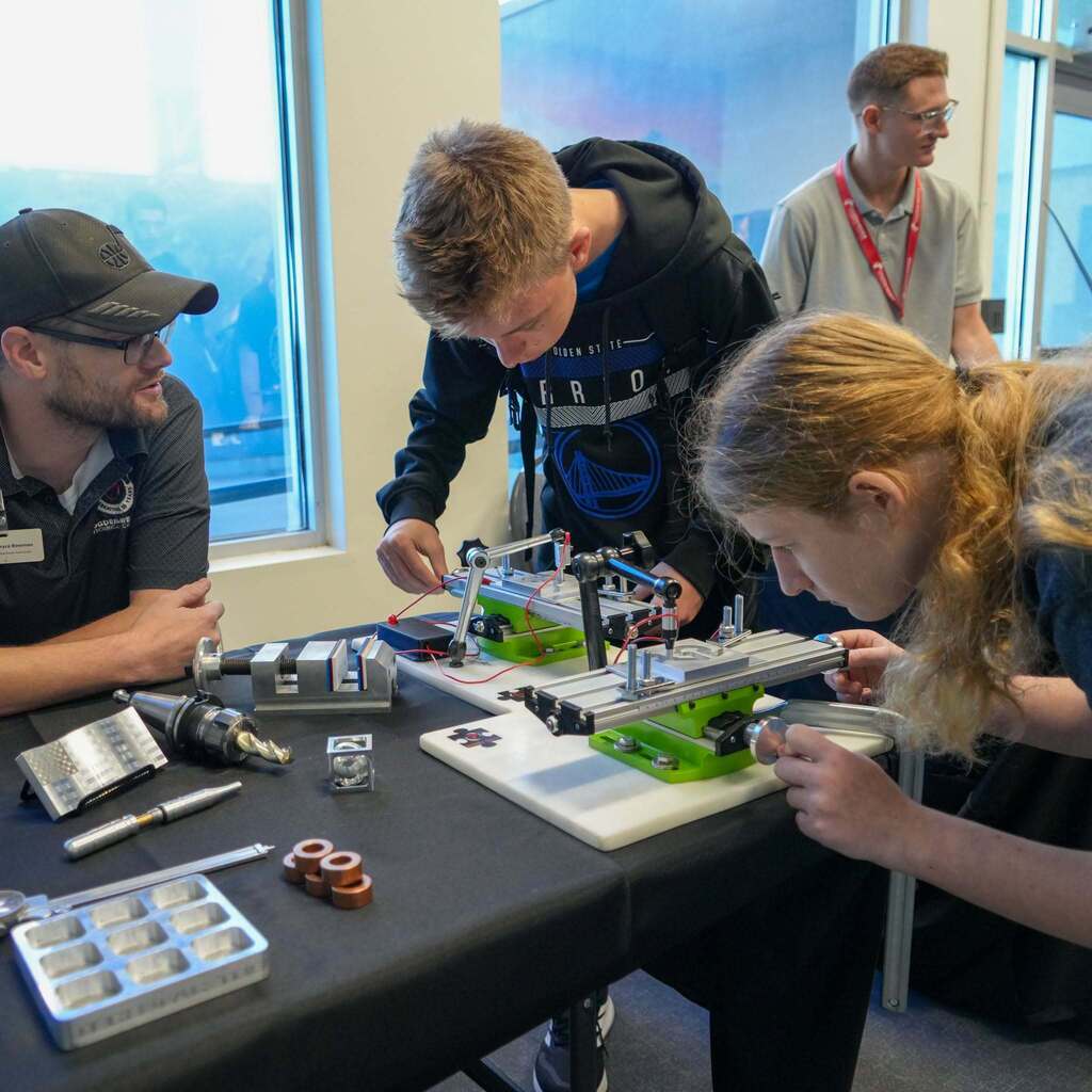 Image 6: Students leaning over a table examining custom-machined mechanical assemblies and precision tools guided by a technical instructor.