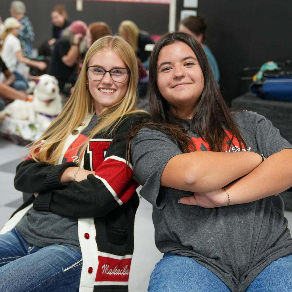 Image 4: Two high school female students sitting side-by-side with arms crossed, smiling at the camera during a school event. One wears a letterman cardigan, and the other wears a grey shirt.