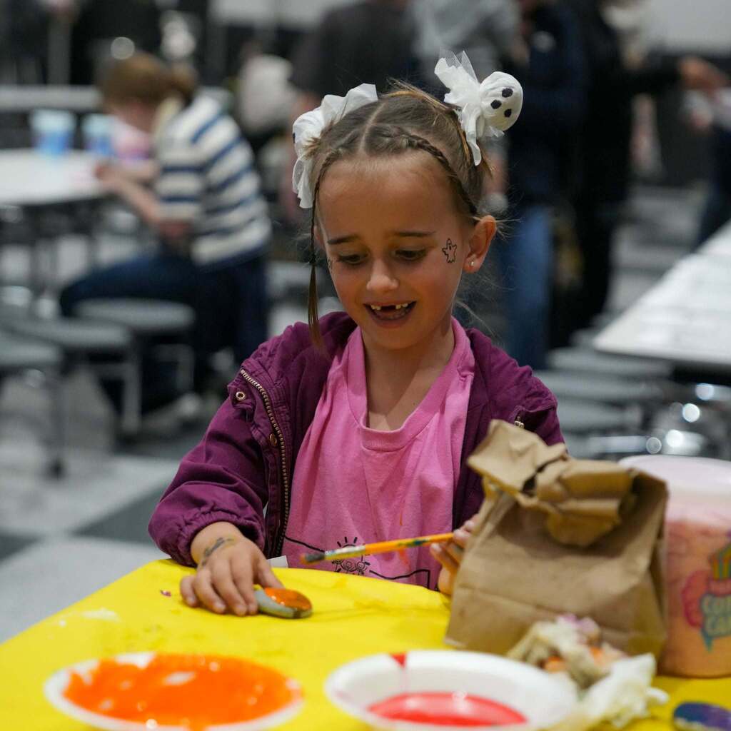 Image 3: A young girl with ghost hair clips and subtle face paint smiling as she paints a rock at a yellow table during an activity.