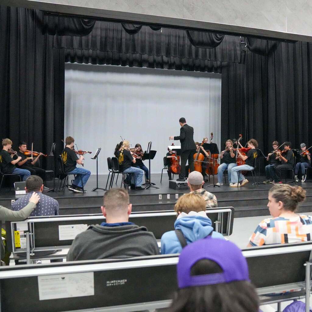 Image 9: A student orchestra performing on stage under a conductor's lead, viewed from the perspective of an audience seated in auditorium chairs.