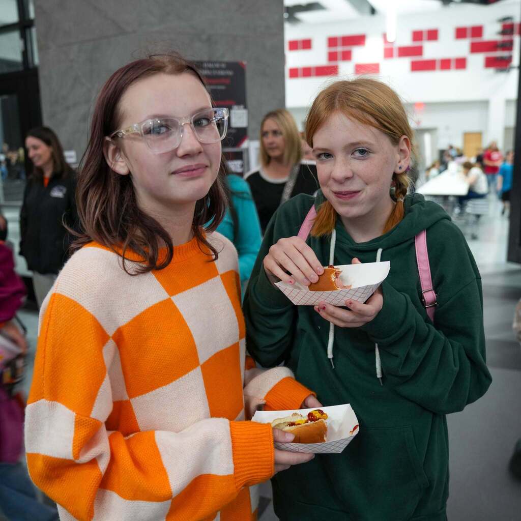 Image 5: Two high school students standing indoors, smiling while holding paper trays with hot dogs. One wears an orange and white checkered sweater, and the other wears a green hoodie.