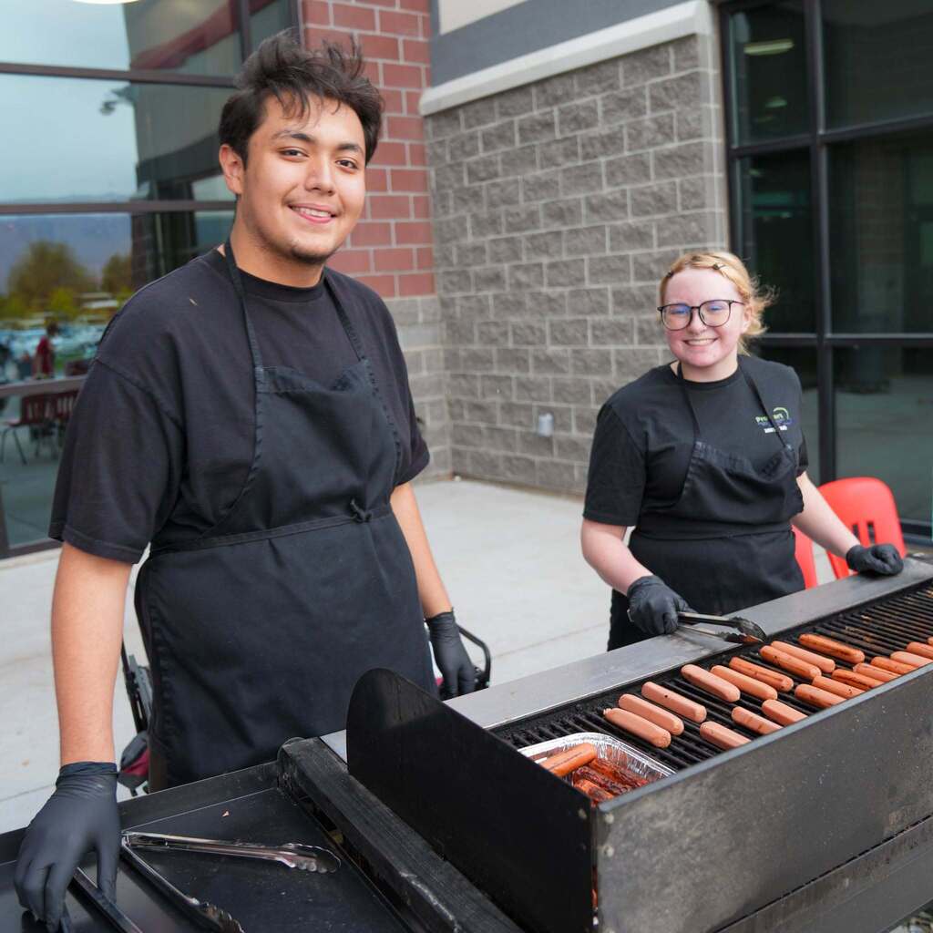 Image 8: Two culinary students wearing black aprons and gloves smiling beside an outdoor grill loaded with hot dogs.